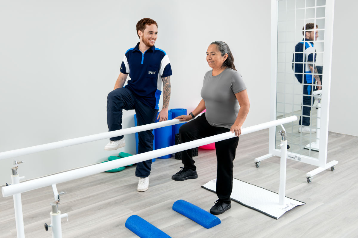 estudiante de fisioterapia de ipeth acompaña a una paciente adulta durante una sesión de rehabilitación física en barras paralelas
