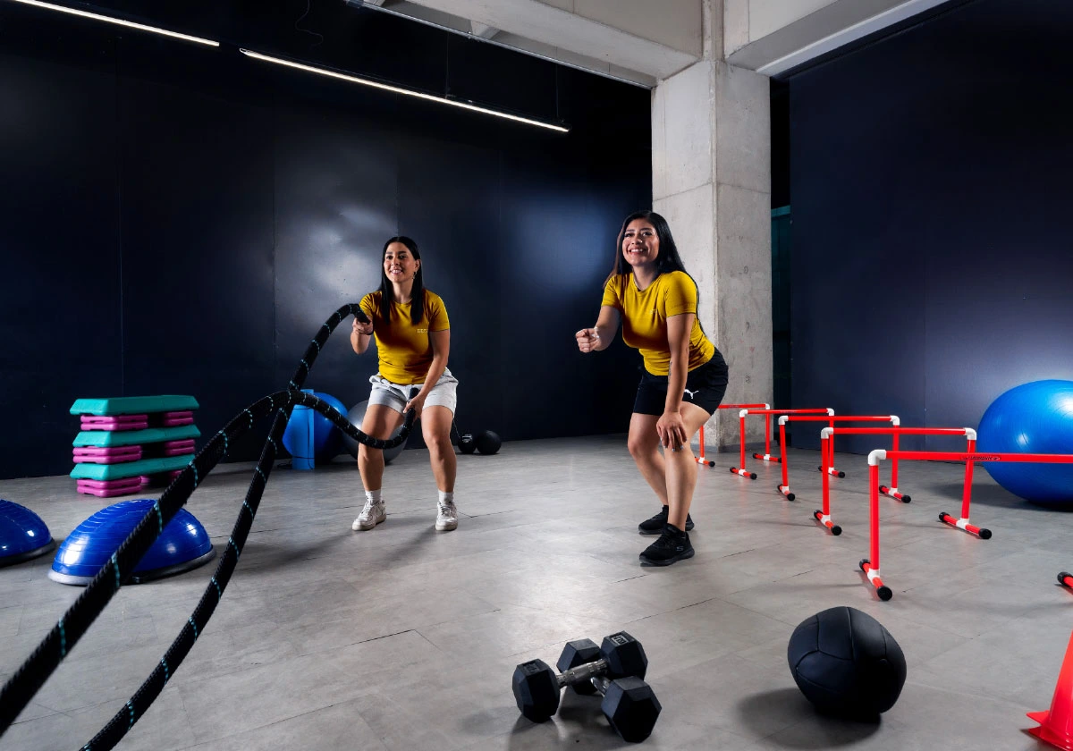dos estudiantes de la licenciatura en ciencias del deporte de ipeth realizan ejercicios funcionales en el área de entrenamiento, utilizando cuerdas de battle rope, vallas, balones y equipo especializado